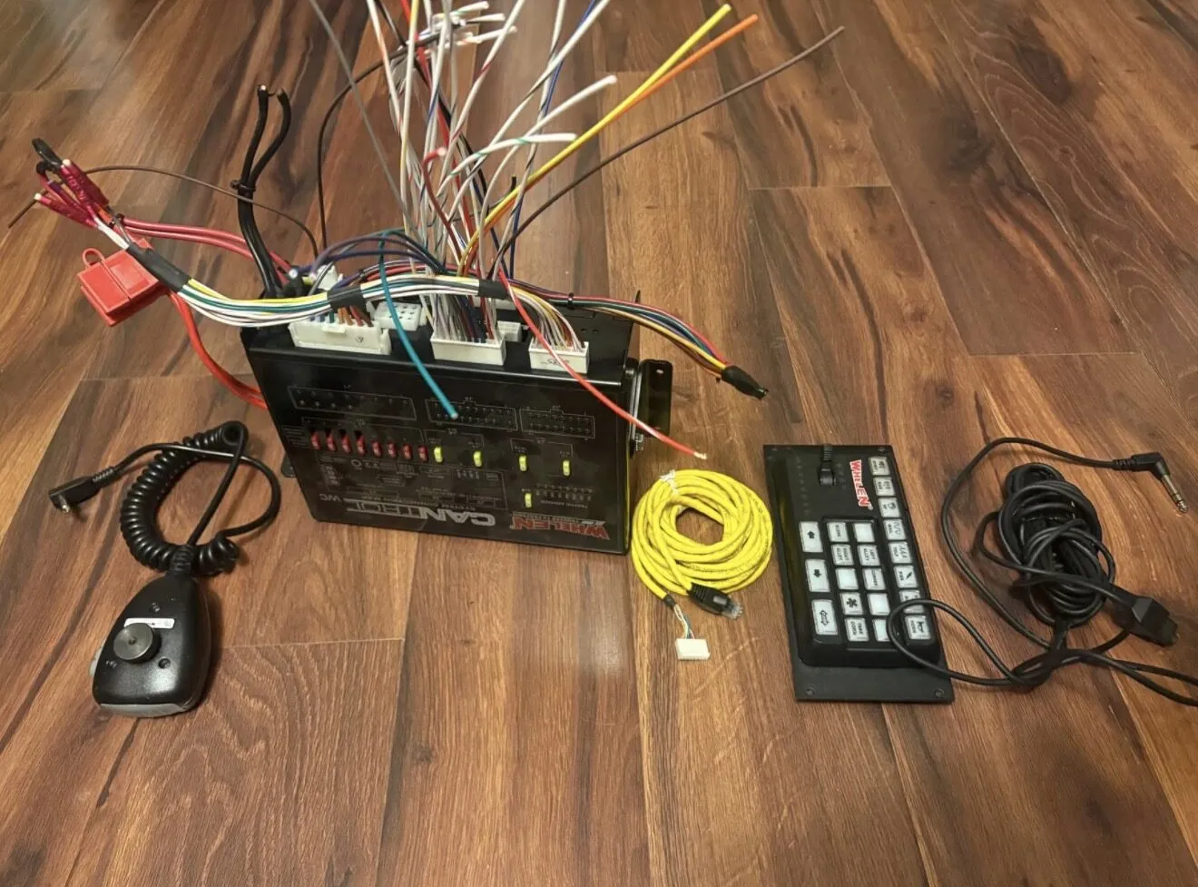 The used Whelen CanTrol WC set on a wood floor: amplifier module with its full wiring harness, the 21-button control head, the yellow head cable and the PA microphone.