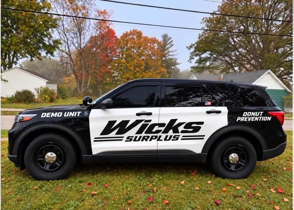 Side view of the Wicks Surplus demo unit: a black Ford Police Interceptor Utility with white doors carrying the WICKS SURPLUS wordmark, DEMO UNIT on the front fender and DONUT PREVENTION on the rear quarter.