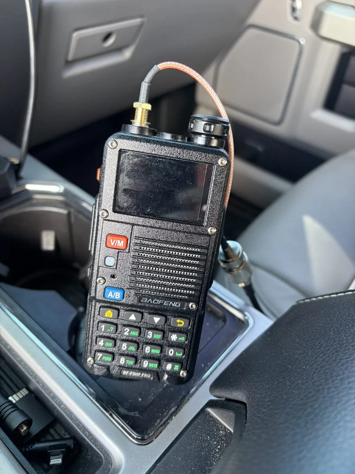 BaoFeng handheld radio standing in the console tray with a short tan SMA jumper running from its antenna port to the coax.